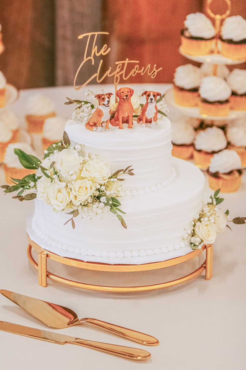 Elegant white wedding cake decorated with fresh flowers and greenery on gold cake stand with matching utensils.