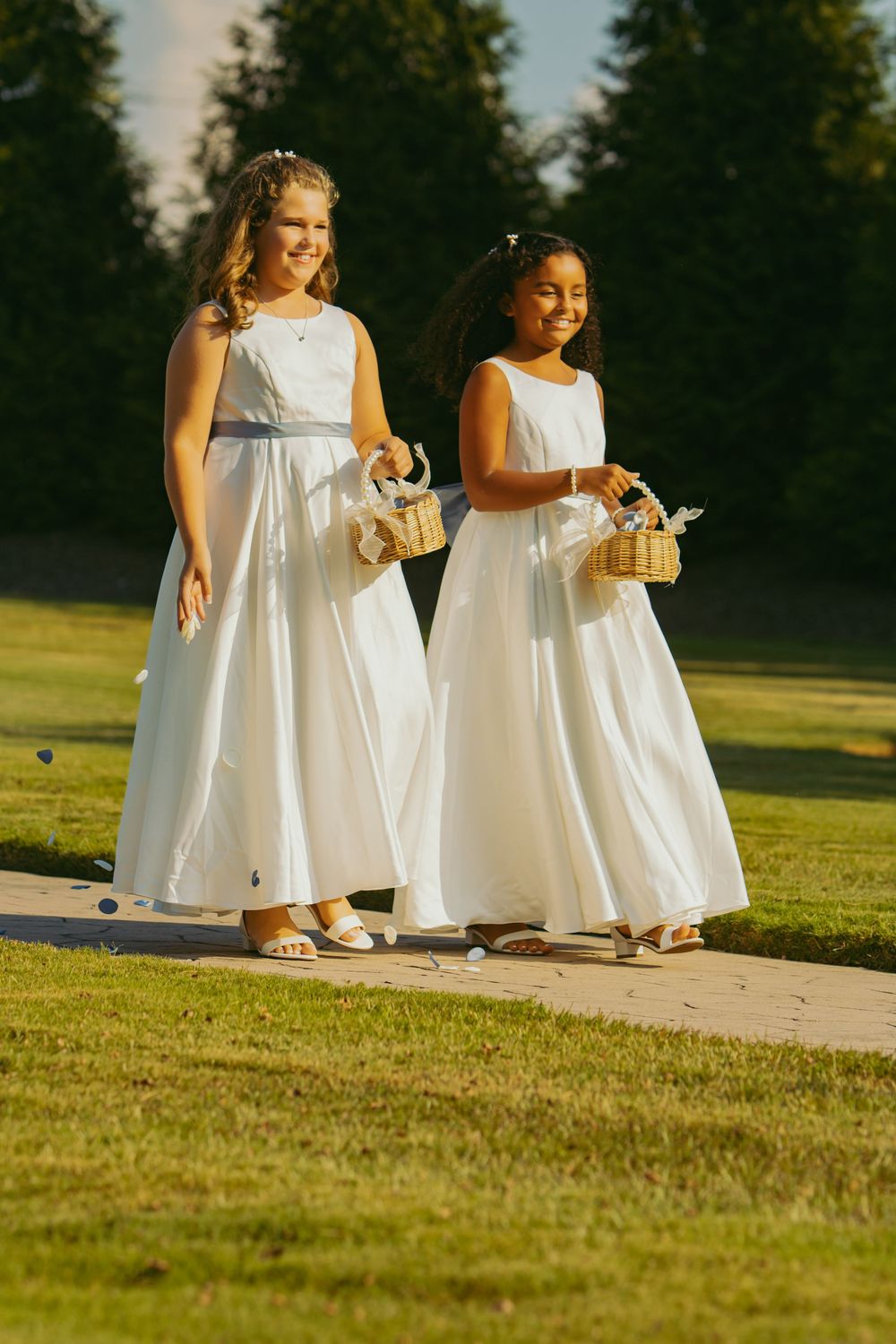 Two flower girls in white dresses walk together on a grassy lawn at sunset carrying small baskets.