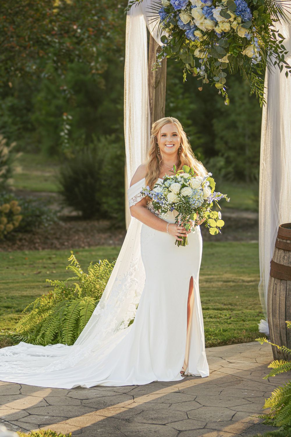 A bride in a flowing white dress poses under a decorated outdoor wedding arch with blue and yellow flowers.
