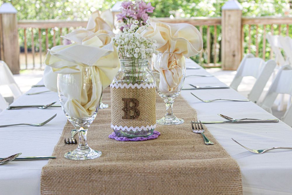 Rustic wedding table setting with burlap runner, mason jar centerpiece with purple flowers, and elegant place settings.