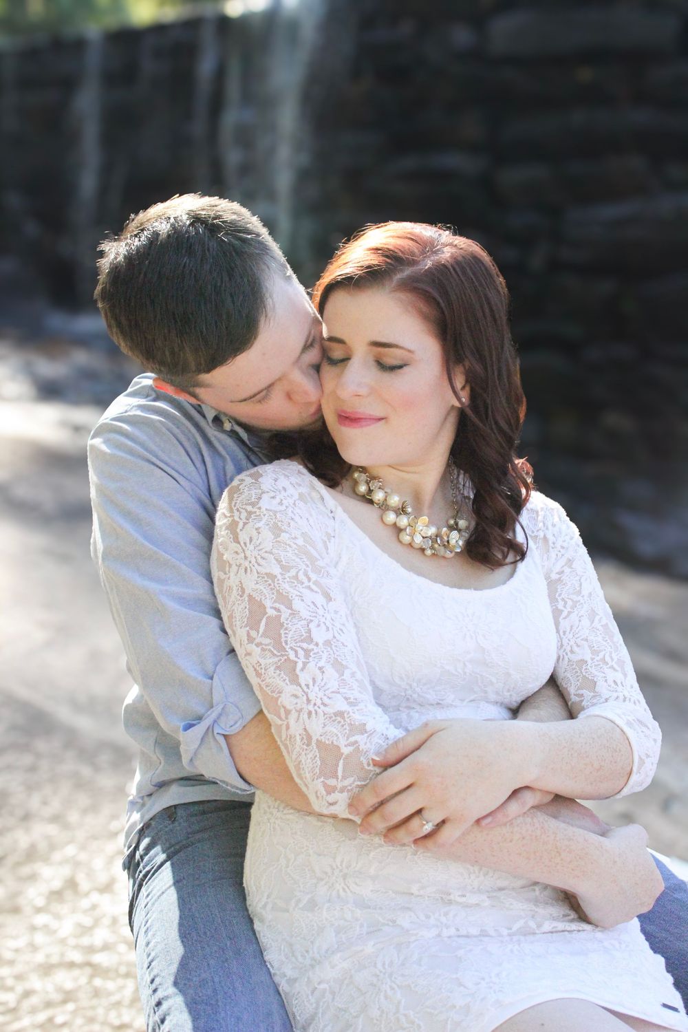 A couple embraces tenderly in an outdoor setting wearing white lace and gray as sunlight filters through trees.