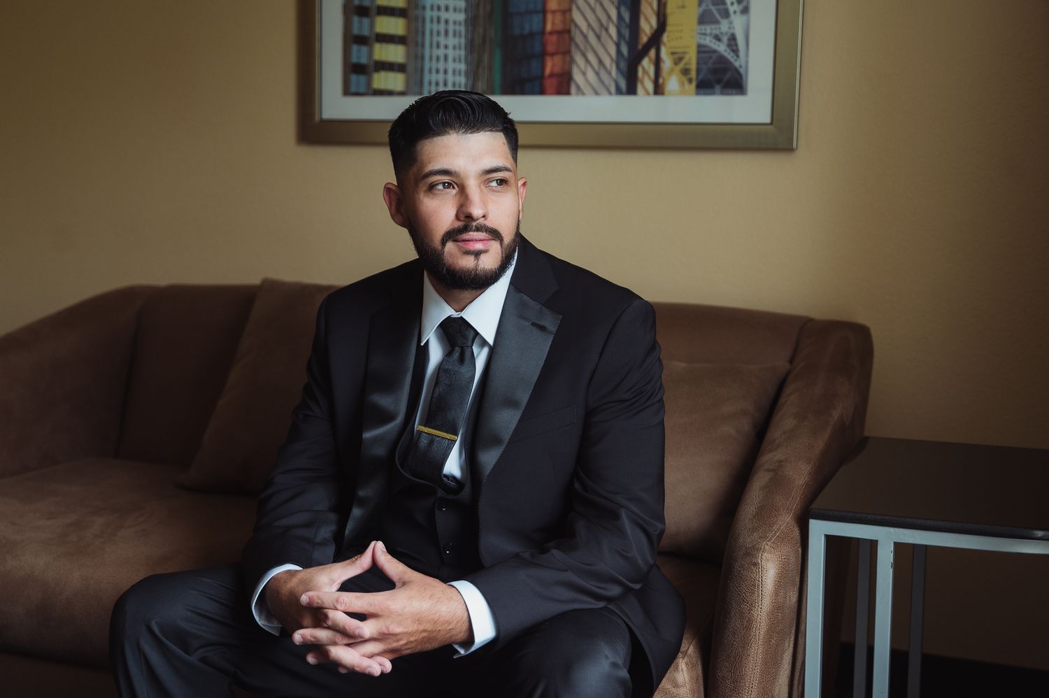 Professional wearing a dark suit sits on brown leather couch while holding smartphone in office setting.