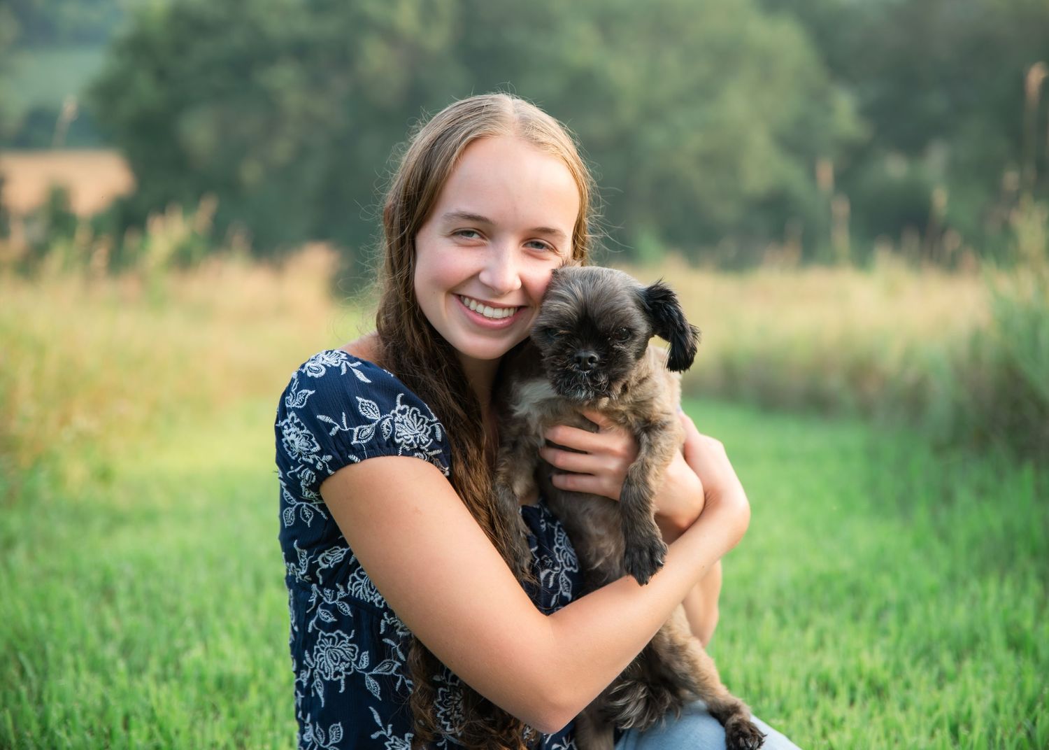 A person in a floral dress holds and cuddles with a small black puppy in a grassy outdoor setting.