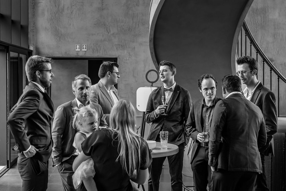 A black and white candid photo shows a group of formally dressed people engaged in conversation under an arched doorway.