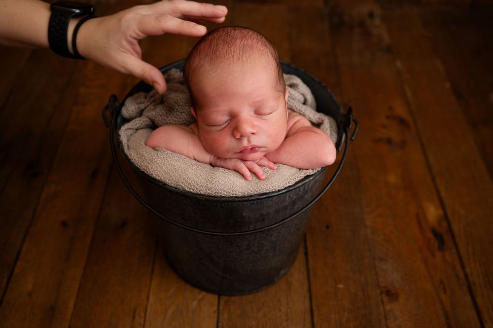 A sleeping newborn baby nestled in a dark bucket with soft padding on a wooden floor, with a gentle hand nearby.