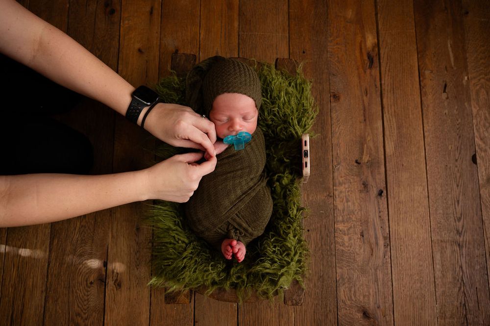 Hands holding a pacifier over a newborn swaddled in olive green on a grassy bed against wooden floor background.