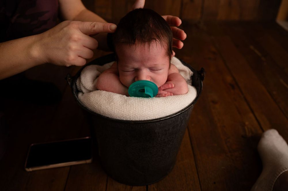 Newborn baby rests peacefully in a cushioned black container with a teal pacifier.