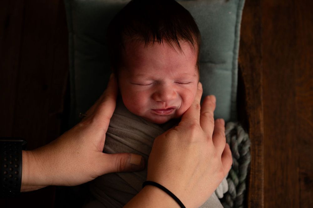A newborn baby smiles while being cradled in soft grey fabric against a dark background.