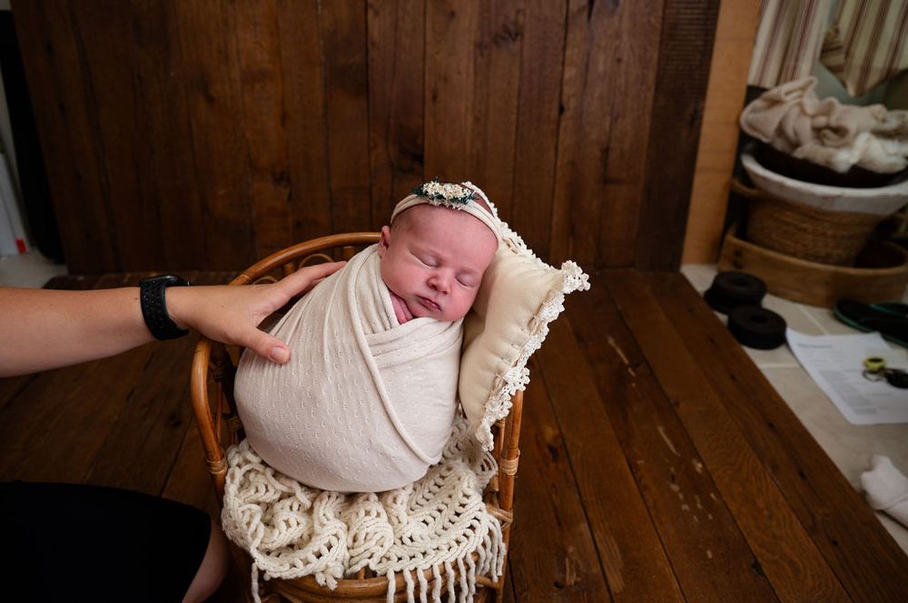 Newborn baby wrapped in cream-colored swaddle rests peacefully in vintage wooden chair with macrame cushioning.