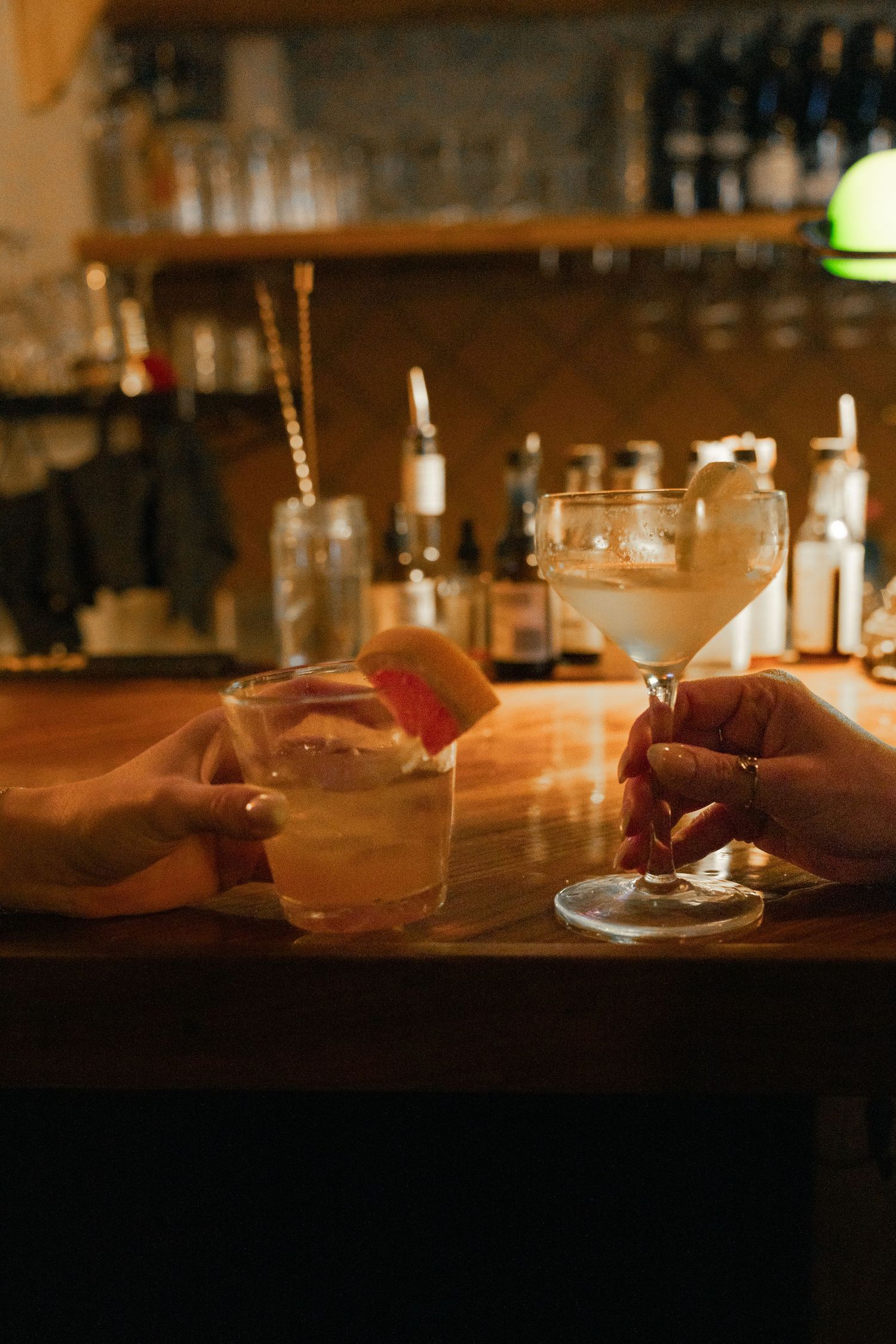 Two hands holding cocktails at a warmly lit bar, one in a rocks glass with citrus garnish, one in a coupe glass.