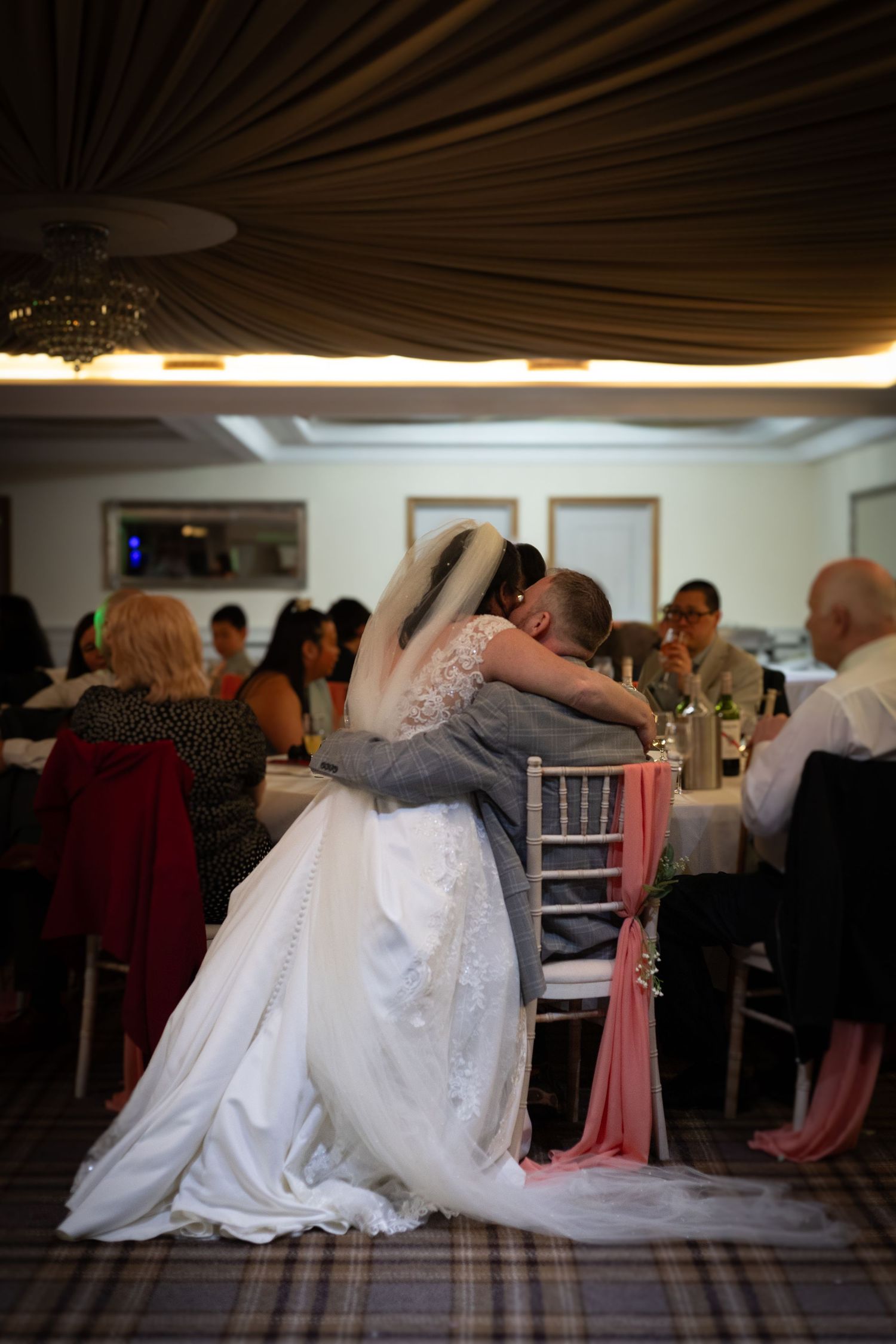 Bride in white gown and veil embraces groom at wedding reception with guests seated at tables in elegant venue.