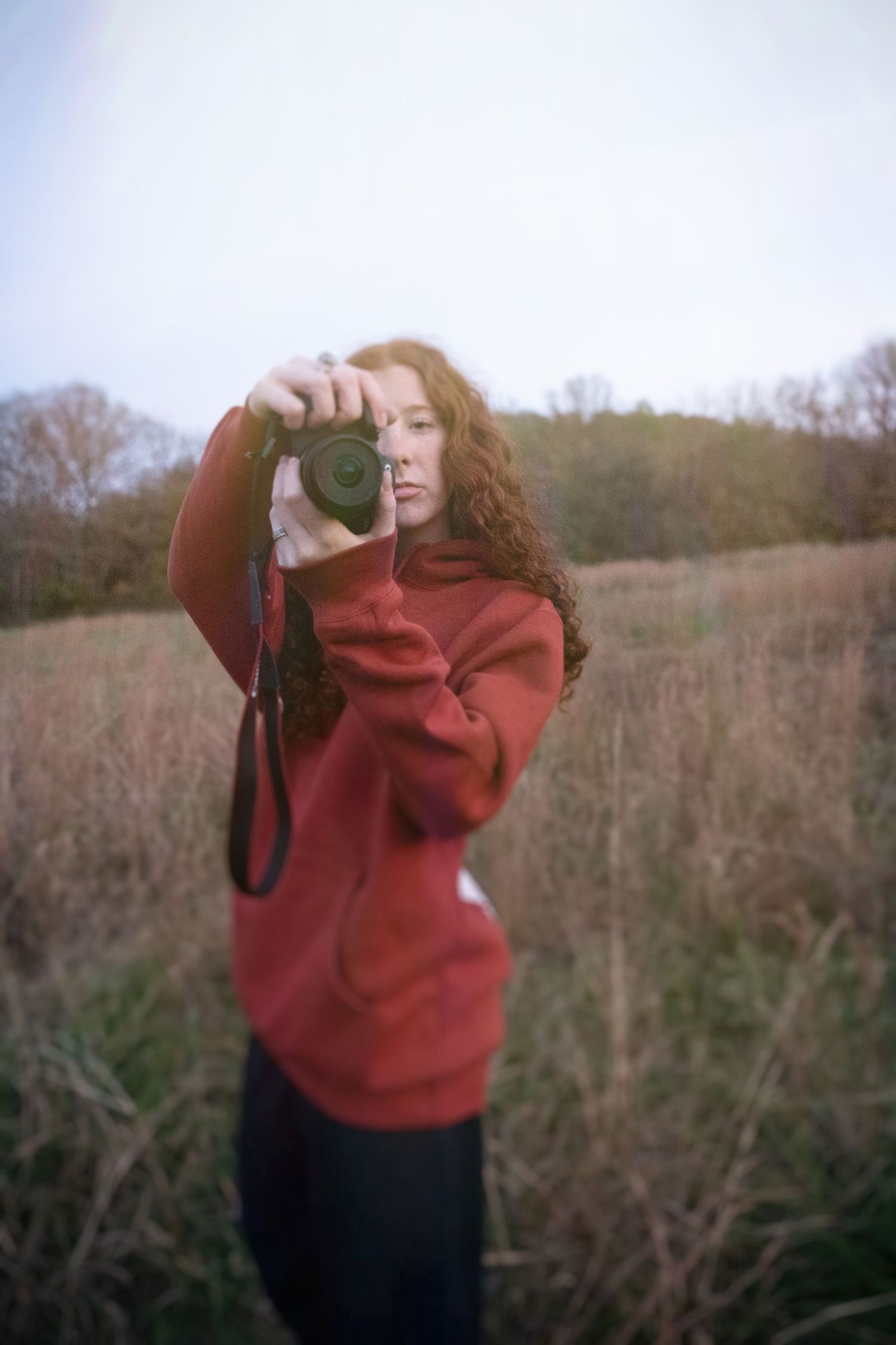 A photographer in a red sweater takes pictures against a backdrop of bare winter trees and bushes.