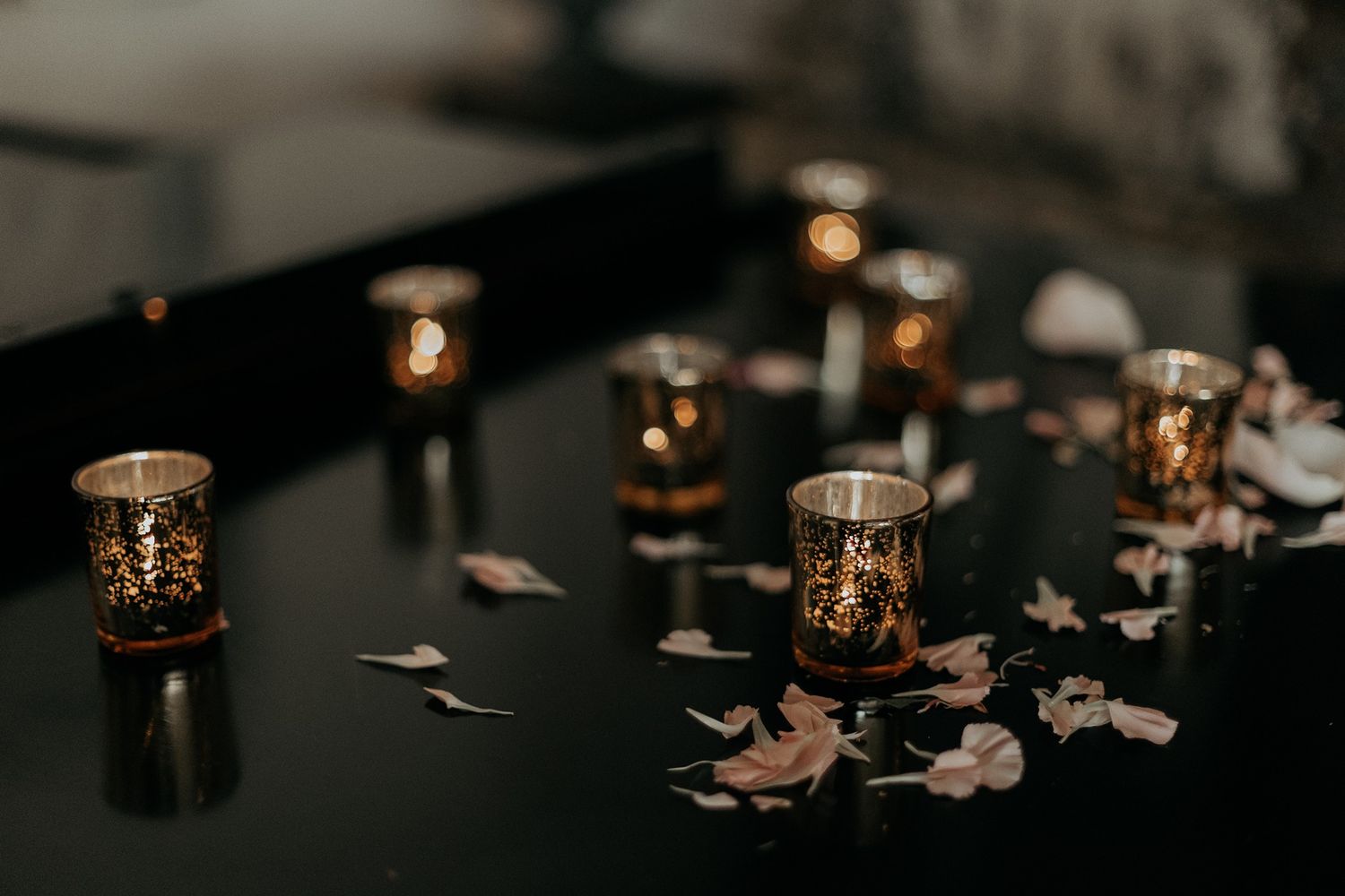 Gold mercury glass votive candles with scattered flower petals on a dark reflective surface.