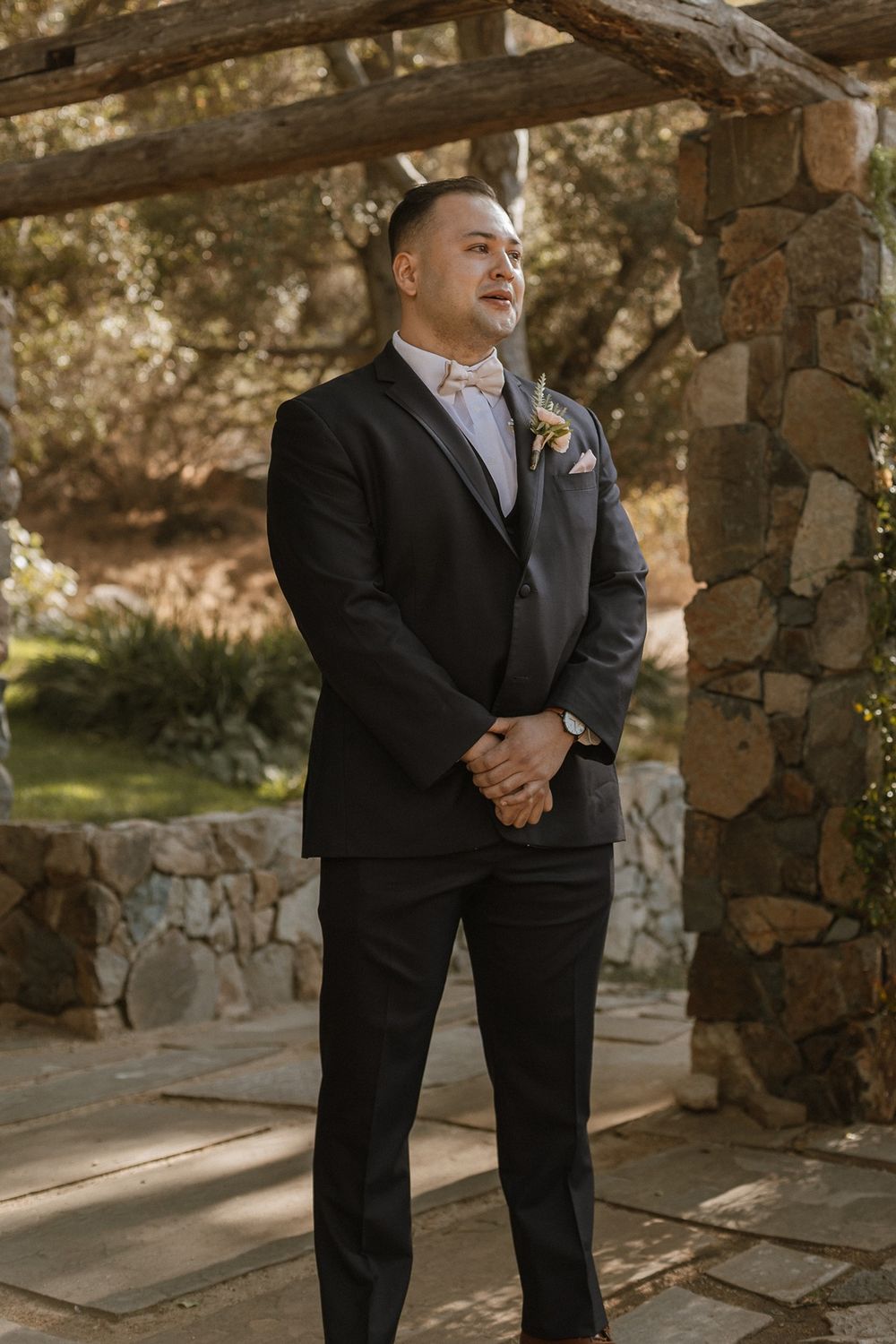 Groom in black tuxedo with floral boutonniere stands under rustic wooden pergola at outdoor wedding venue.