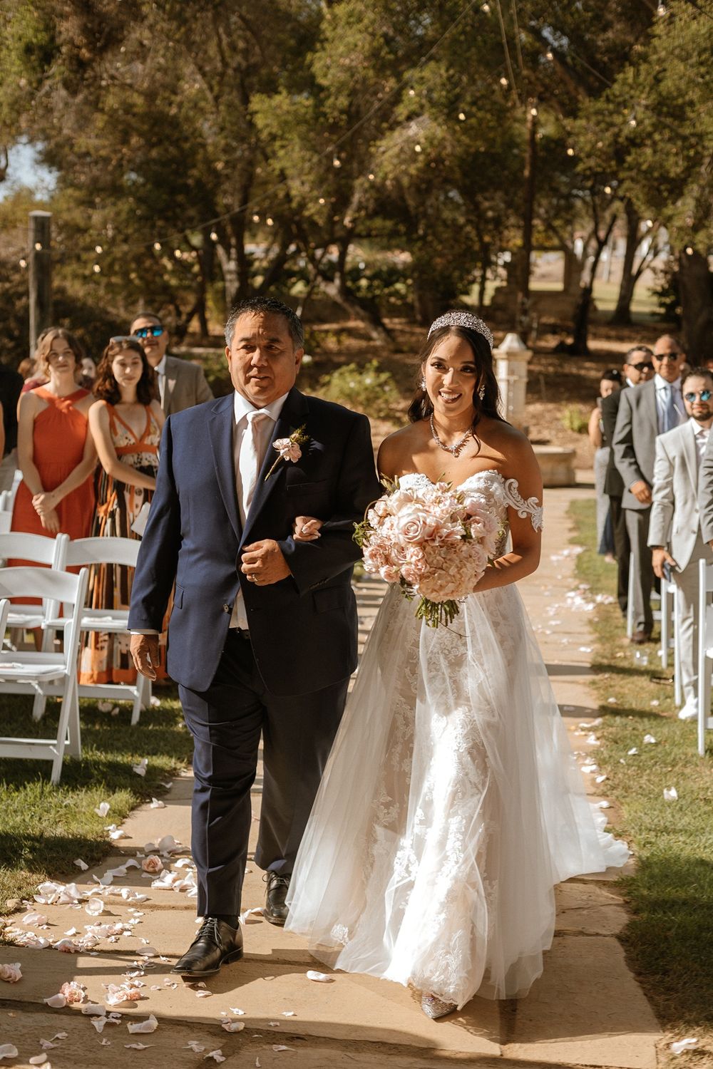 Bride in lace gown walks down aisle with escort at sunny outdoor wedding ceremony.