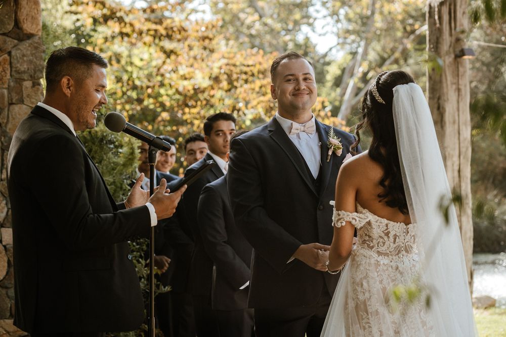 Outdoor wedding ceremony with groom smiling at bride in lace dress, officiant speaking at microphone, stone backdrop.