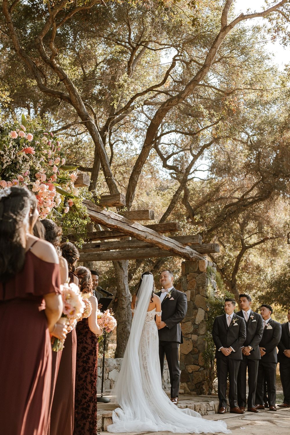 Outdoor wedding ceremony under rustic wooden pergola with bride in lace gown, groom, bridesmaids in rust dresses, and groomsmen.