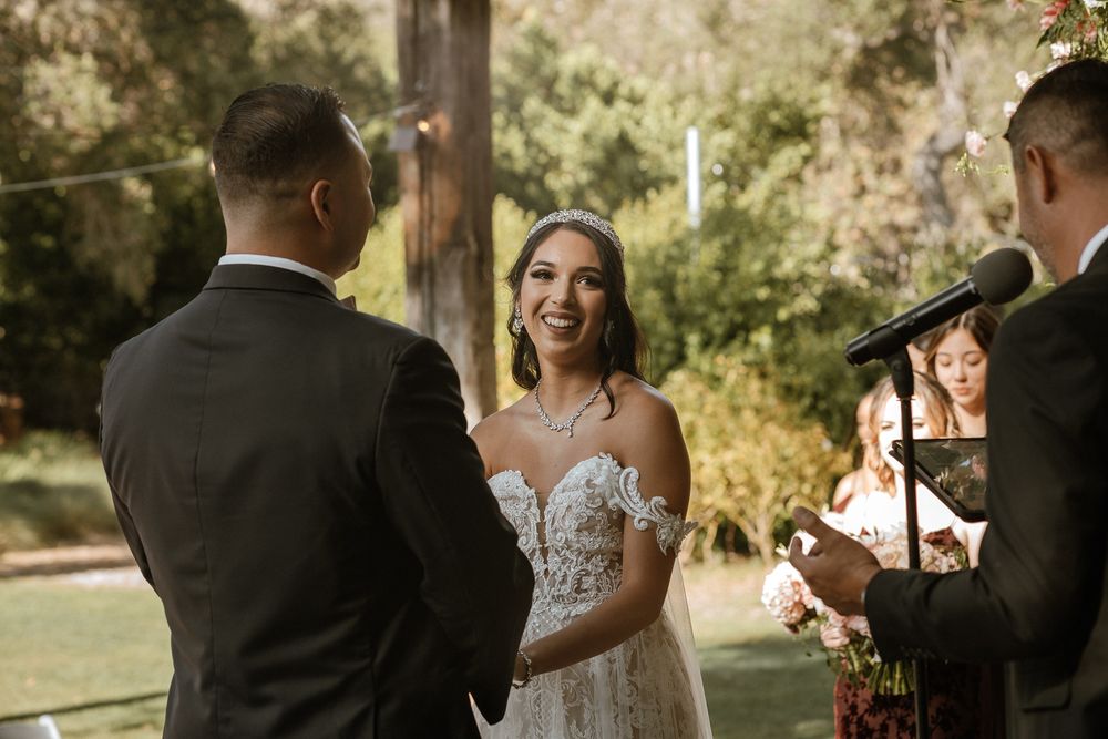 Bride smiling joyfully at groom during outdoor wedding ceremony with officiant and lush green background.