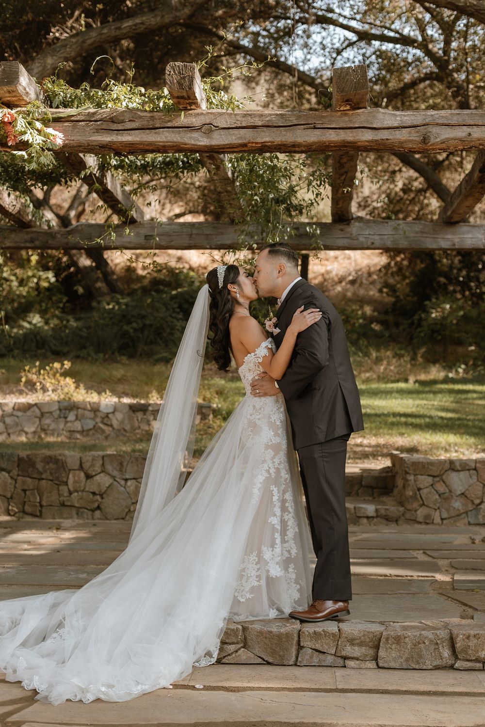 Bride and groom share first kiss under rustic wooden trellis at outdoor stone wedding ceremony surrounded by lush greenery.