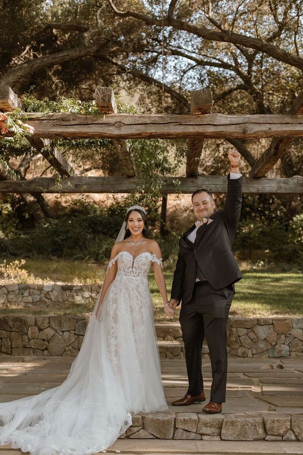Joyful newlyweds celebrate outdoors, bride in lace gown and groom in dark suit raising hands under rustic wooden pergola.