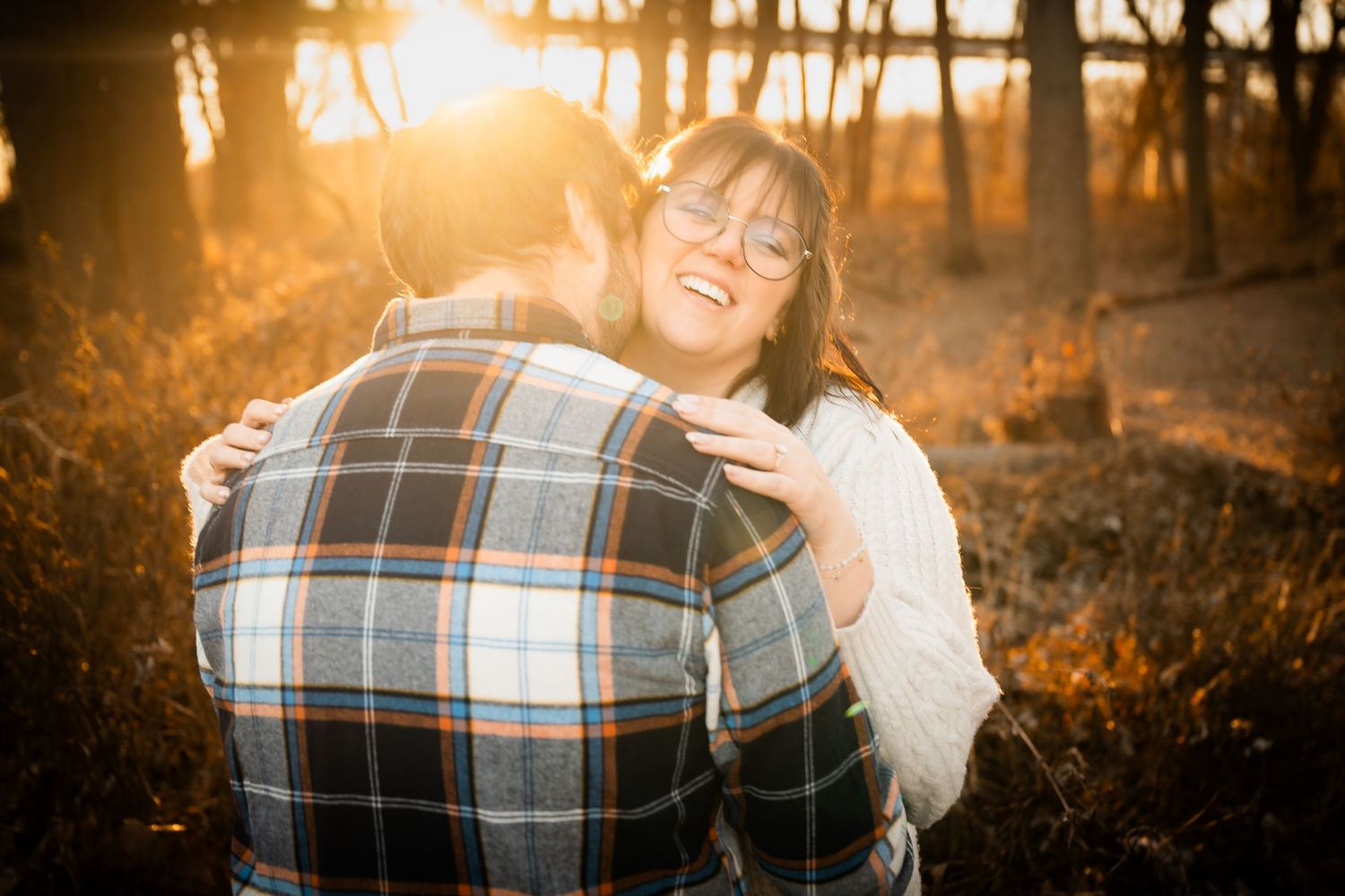 Couple embracing at golden hour sunset in autumn forest, woman smiling joyfully in glasses and white sweater.