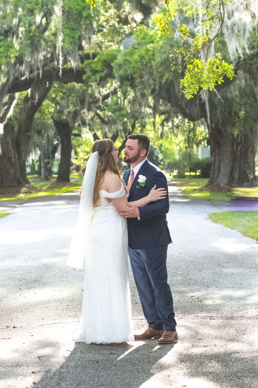 Newlywed couple shares intimate moment on sunlit garden path under moss-draped oak trees.