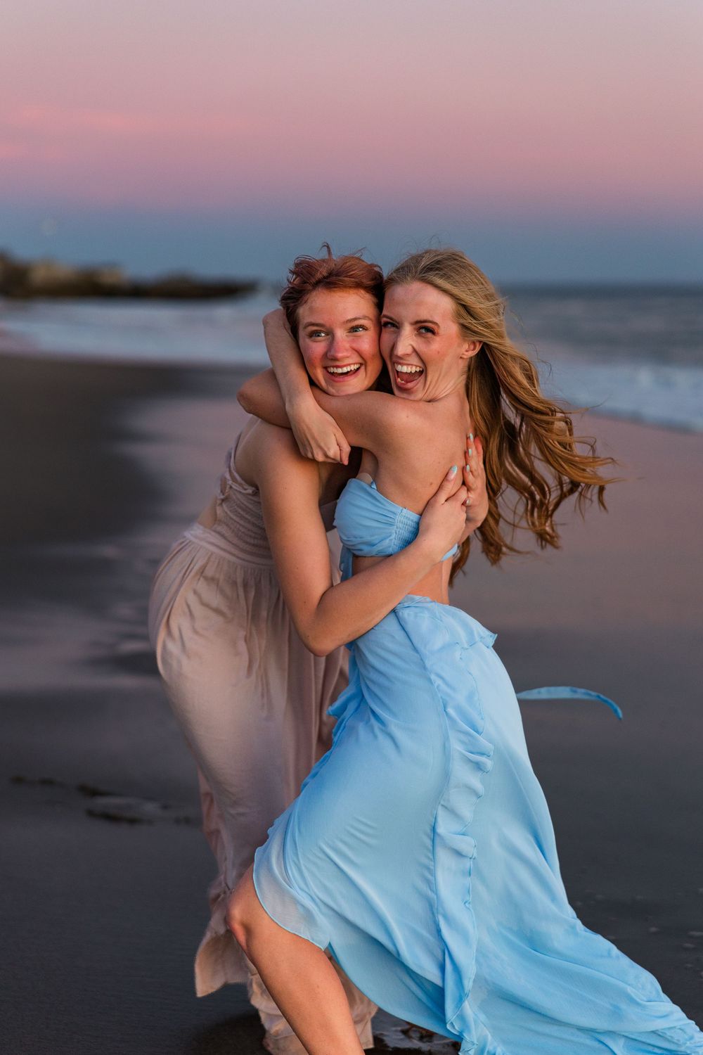 Two friends in flowing dresses laugh and embrace on a beach at sunset with pink and blue sky in the background.