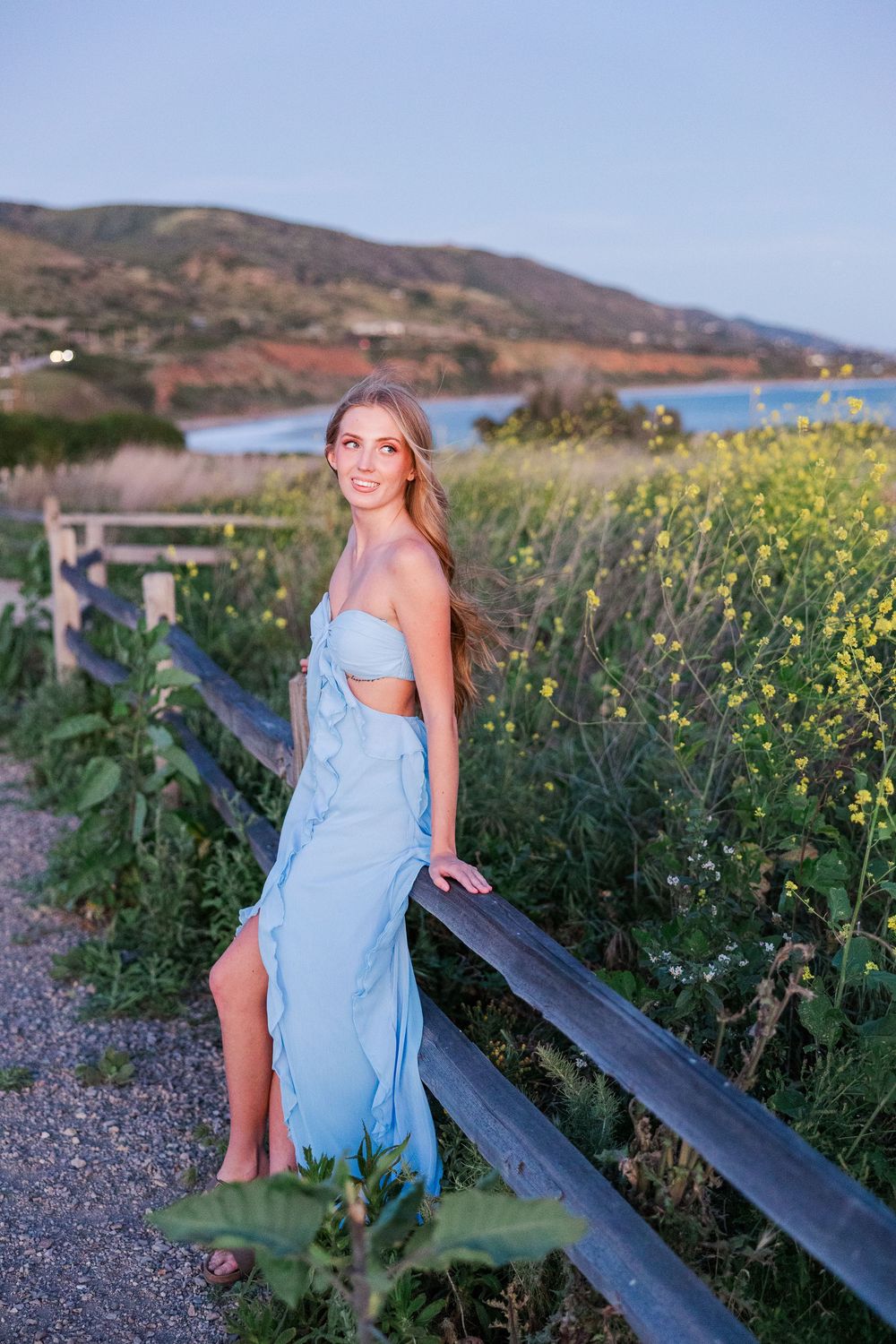 Woman in light blue dress poses in field of wildflowers with mountains and coastline in background at sunset.
