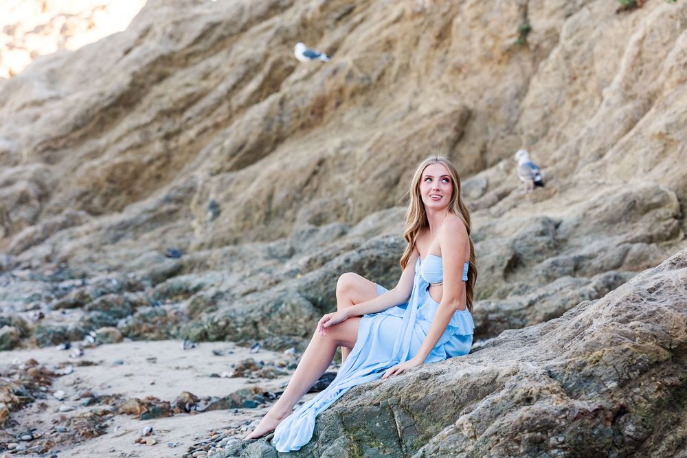 A person in a light blue dress poses on rocky coastal cliffs during sunset creating a dreamy seaside portrait.