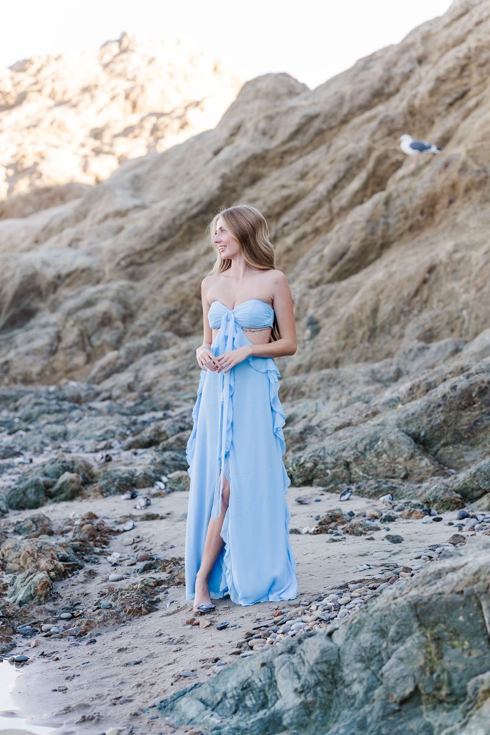 Person in flowing light blue dress sits gracefully on rocky beach at sunset with cliffs in background.