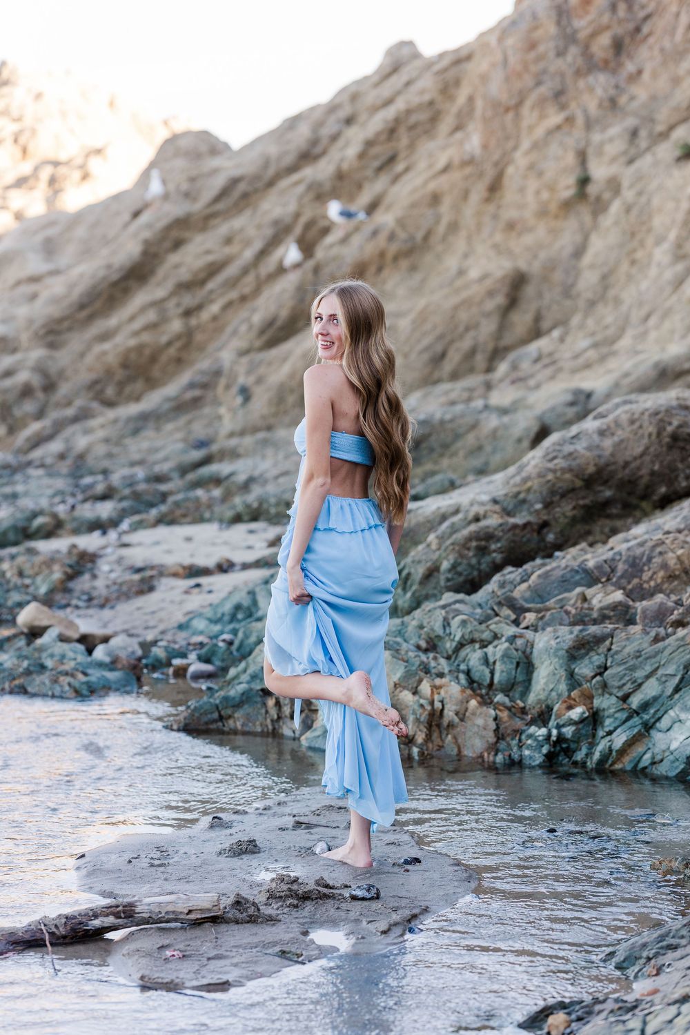 A person in a light blue dress walks gracefully along rocky coastal terrain at sunset.