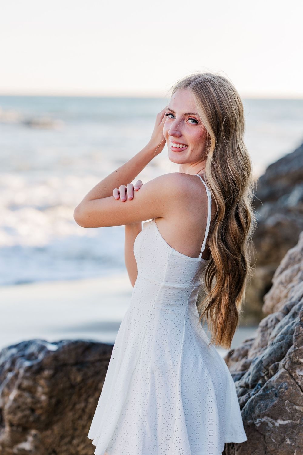 A series of beachside portraits featuring someone in a white eyelet dress posing against a coastal backdrop at sunset.