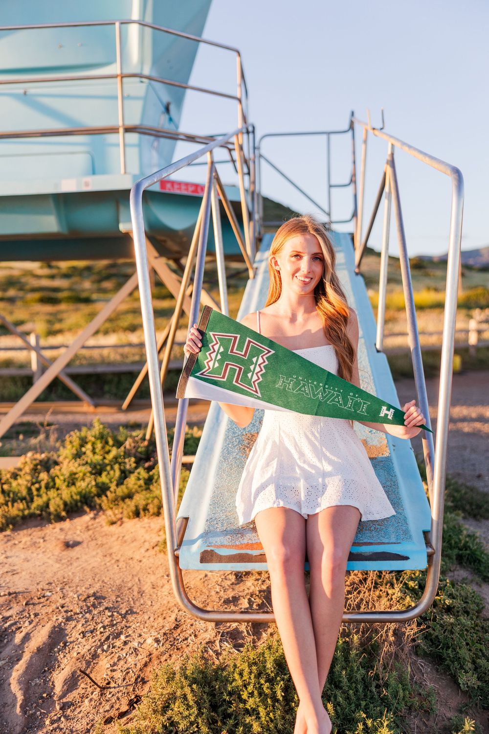Woman in white dress holds green pennant flag while sitting on lifeguard tower at beach.