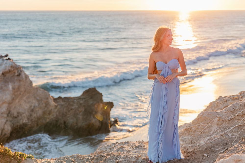Silhouetted figure in flowing blue dress stands on rocky beach at sunset with ocean waves crashing in background.