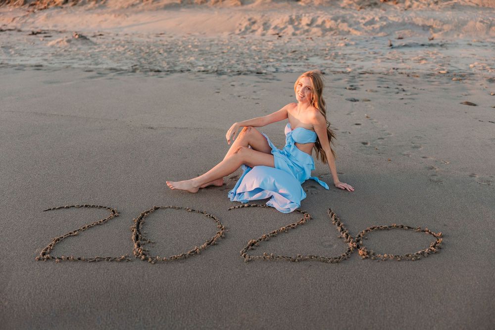 Someone in a blue dress sits on a sandy beach next to "2022" written in the sand at sunset.