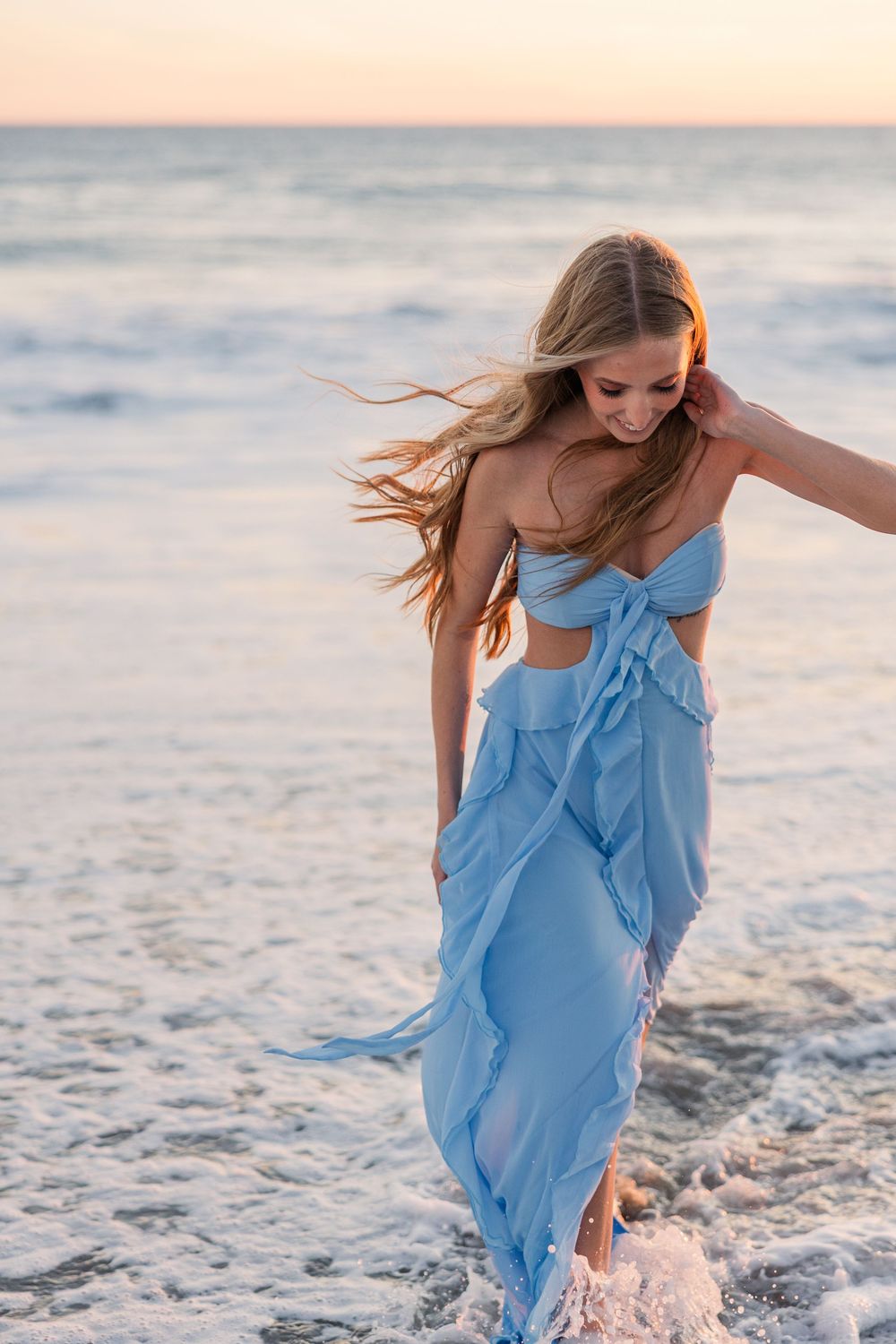 A woman in a flowing light blue dress dances gracefully along the beach at sunset as ocean waves crash behind her.