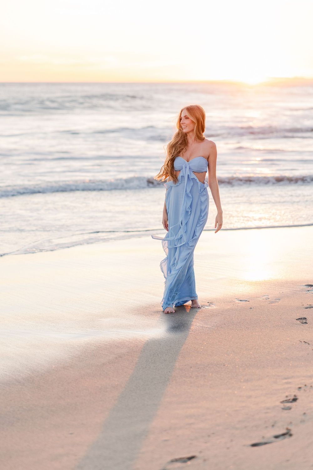 A person in a flowing blue dress walks along a sandy beach at sunset leaving footprints in their wake.