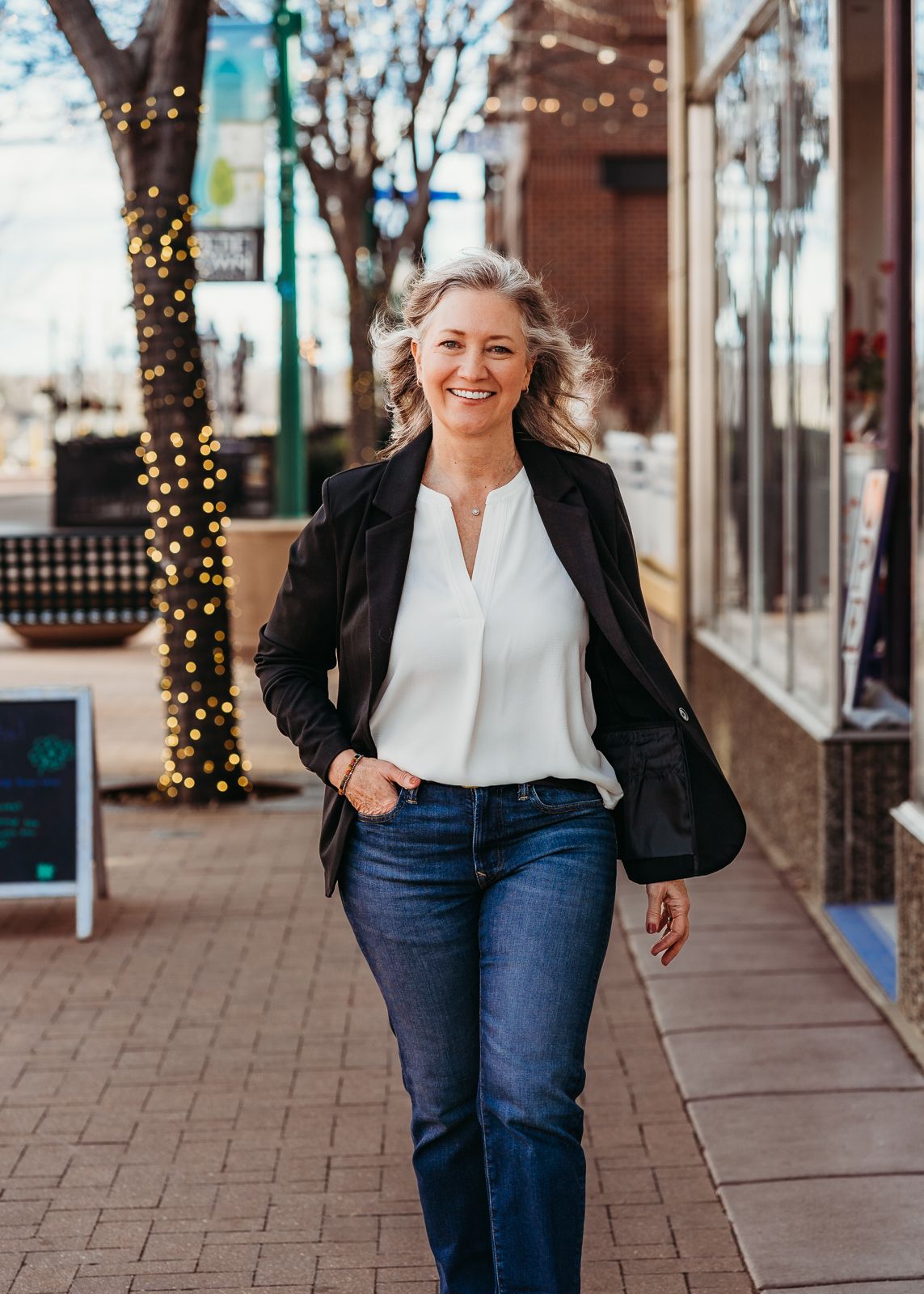 Woman in black blazer and jeans walks along a brick sidewalk with twinkling holiday lights in background.