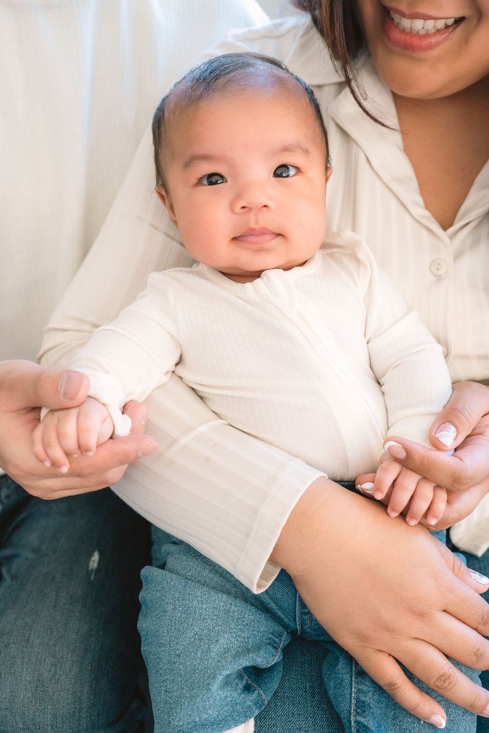 Baby wearing white outfit sits on lap while being held by person in denim and white.
