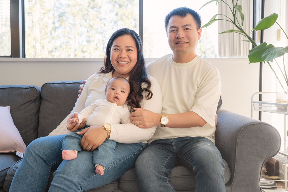 A family relaxes together on a light gray couch in a bright, modern living room with houseplants.