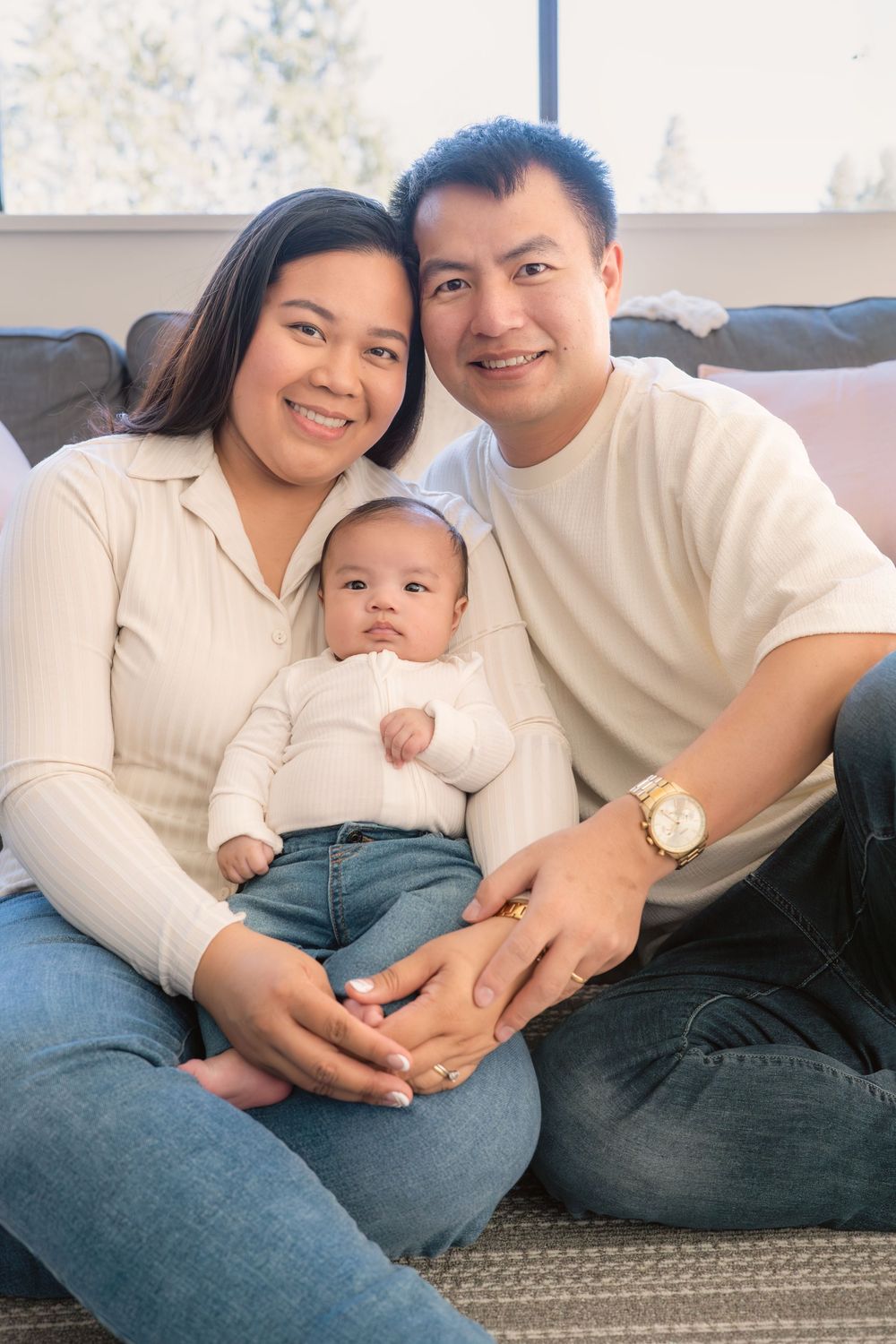 A young family sits together on a couch, with parents in cream colored tops holding their baby who wears white and jeans.