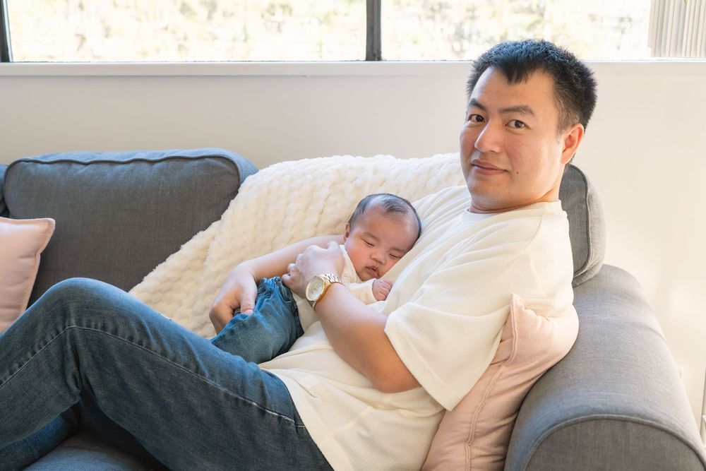 A parent in a white t-shirt relaxes on a gray couch while holding a sleeping baby in blue clothing.