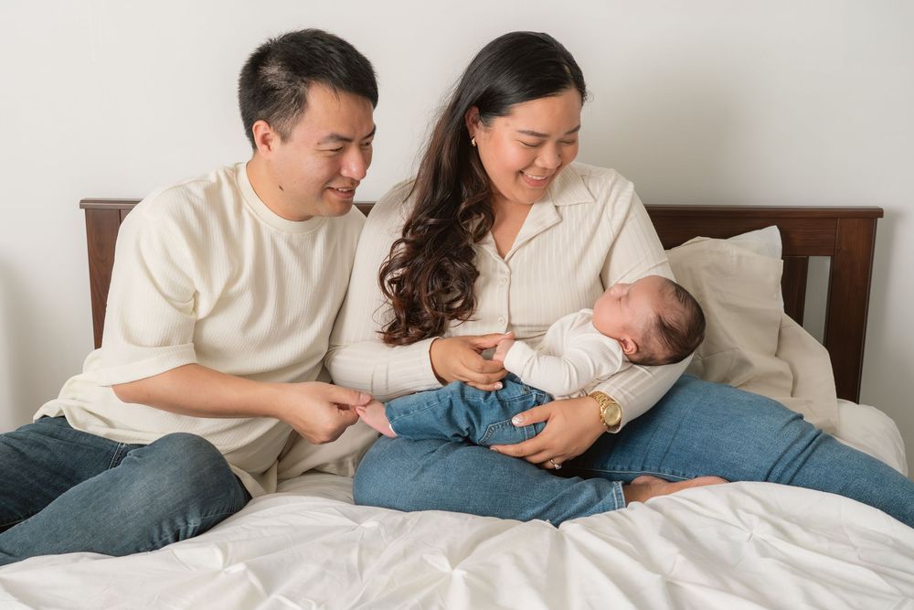 A couple sits on a bed together with their newborn baby, sharing a tender family moment at home.