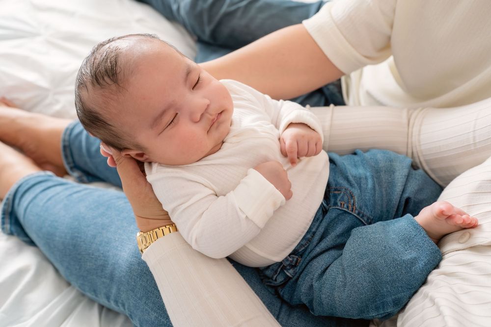 Baby in white sweater and jeans lying on soft white blanket while being comforted by parent's hand.