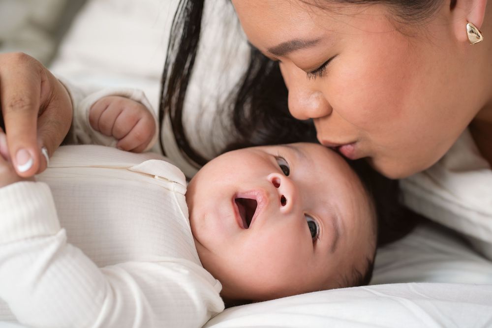 A tender moment captured as a parent kisses their baby who lies on white bedding with a joyful expression.
