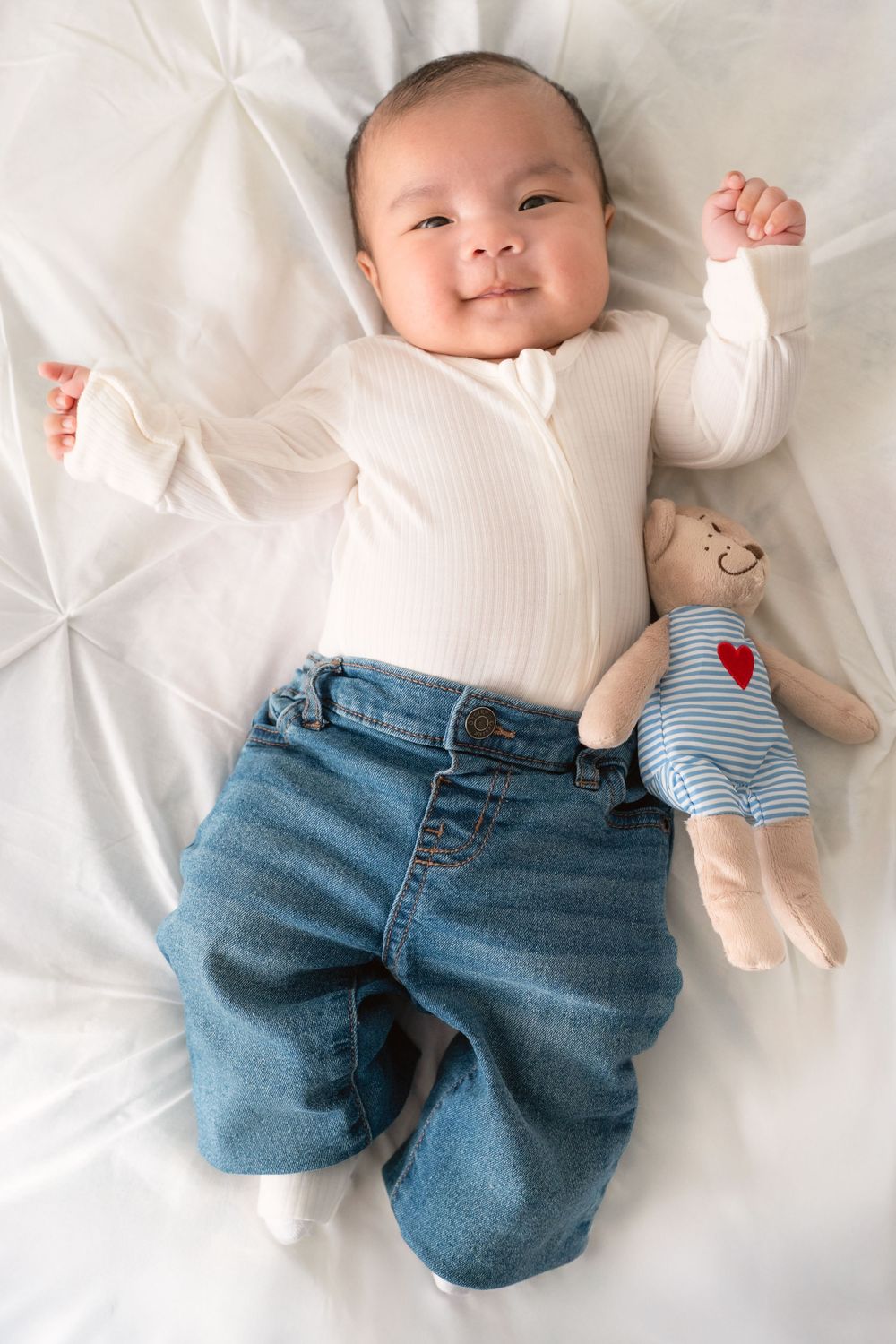 Baby wearing white top and denim jeans lies on white bedding in a series of sweet poses.
