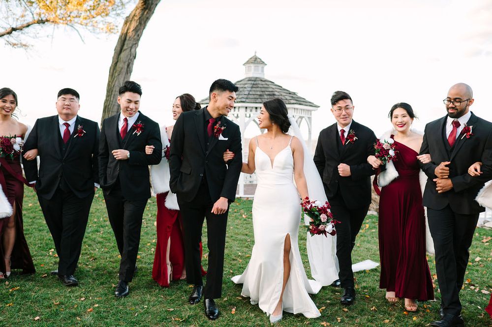 Wedding party in burgundy and black attire poses together outdoors during autumn with fall foliage in background.