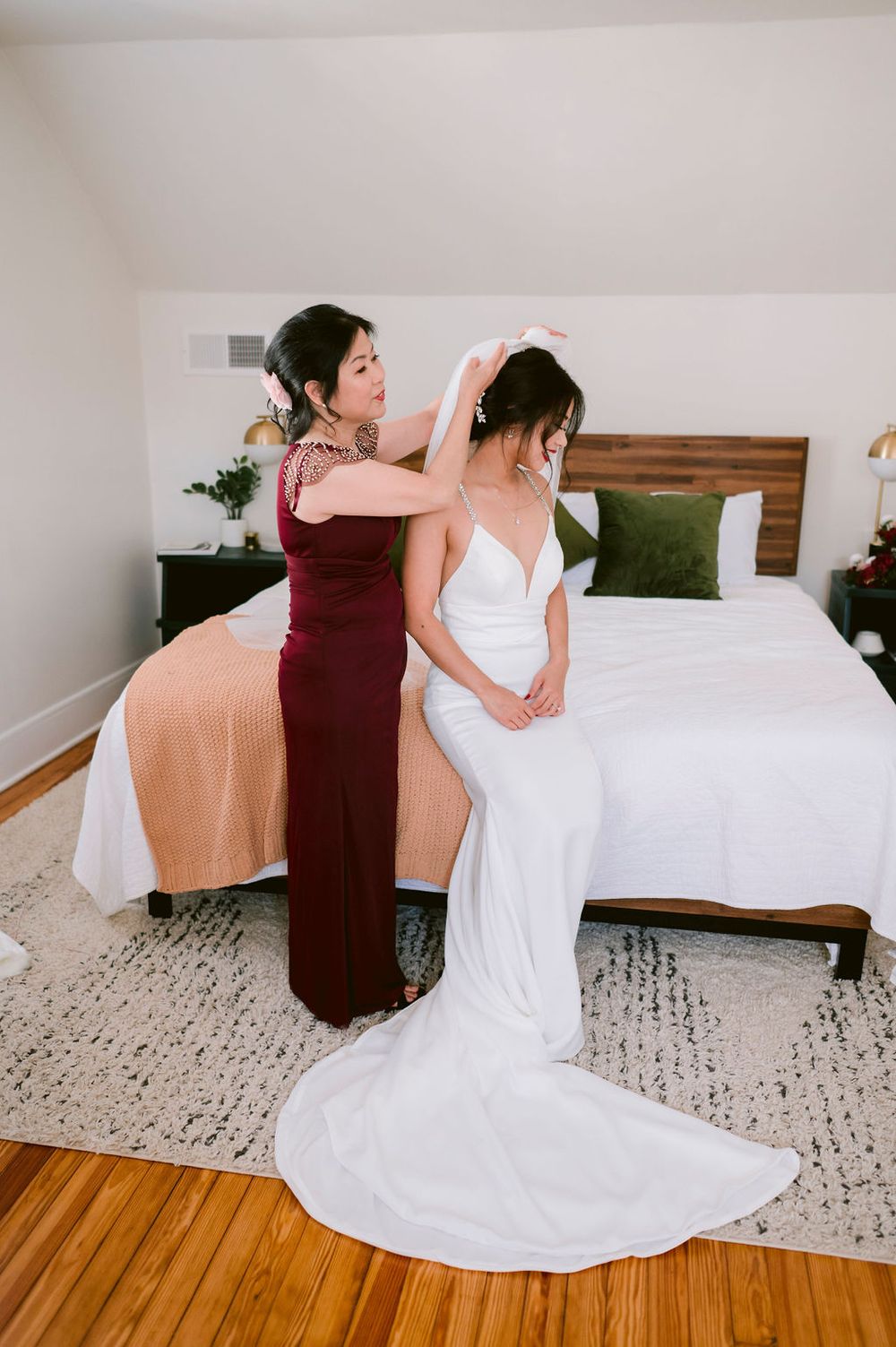 Two people in burgundy and white dresses sitting together on a white bed with a patterned rug below.