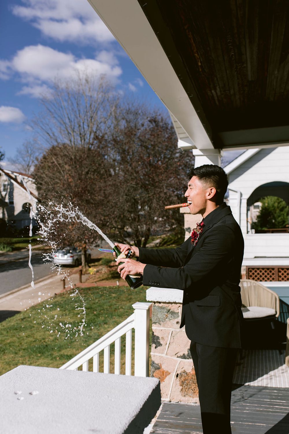 Someone in a dark suit sprays champagne on a porch with white railings on a sunny day.