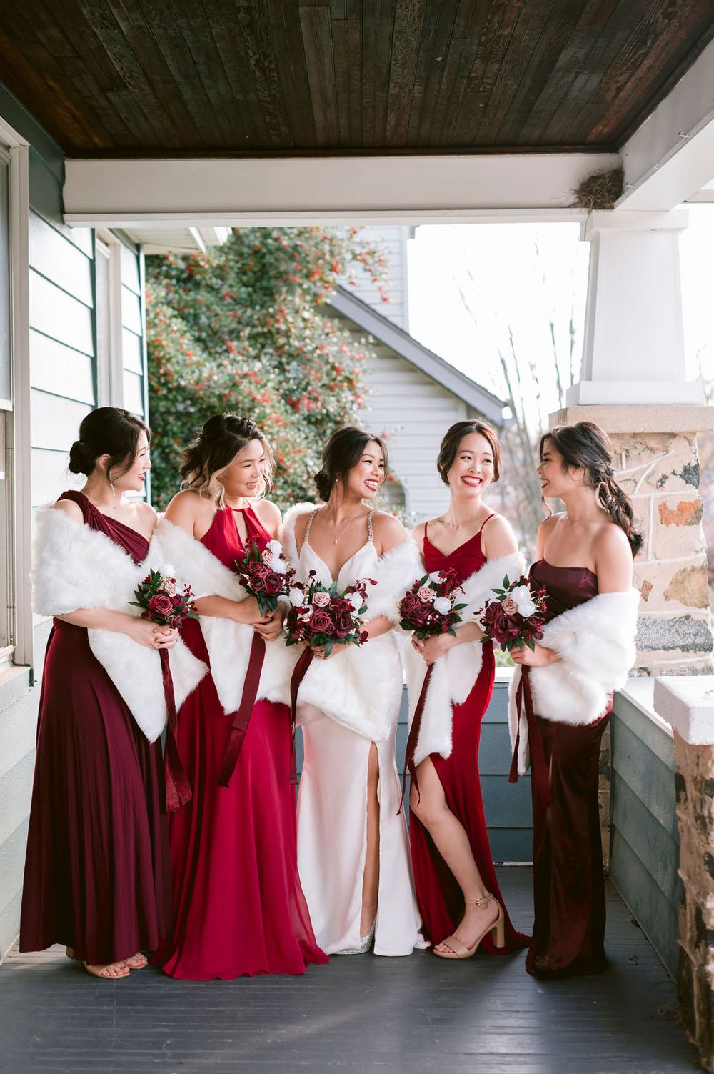Bridesmaids in burgundy dresses share joyful moments on a covered porch during a wedding celebration.